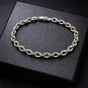 May include: Silver chain bracelet featuring oval links. The bracelet is presented on a black surface. The clasp is visible, and the links have a textured finish. A piece of jewellery.