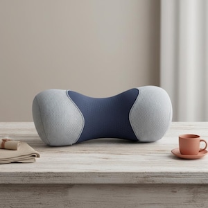 Peut inclure: Un oreiller de cou bleu et gris en forme de sablier. L'oreiller présente une texture en forme de maille sur les extrémités grises et une texture lisse et perforée au centre bleu. Une petite tasse à thé et une soucoupe pêche sont sur une table en bois.
