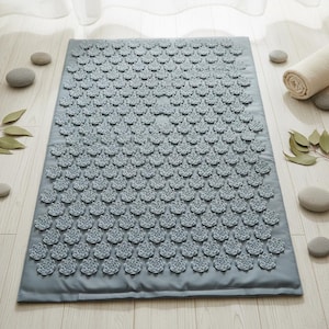 Peut inclure: Un tapis d'acupression rectangulaire gris doté d'une grille dense de points de stimulation en plastique gris clair en forme de lotus couvrant toute la surface. Le tapis présente une bordure en tissu lisse et un motif uniforme de picots.