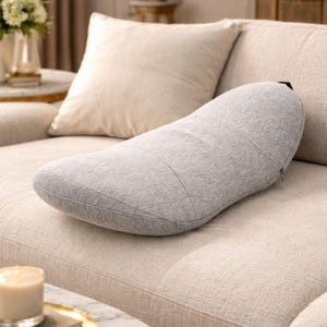 May include: A light grey, kidney-shaped cushion lies on a beige sofa. The cushion has a curved design with stitched detailing and a zip closure. A square cushion and the sofa back are visible in the background.
