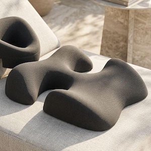 Puede incluir: Dos cojines ergonómicos de color gris oscuro con un diseño curvo único. Los cojines están hechos de una tela texturizada y se colocan sobre una superficie gris claro. Una pequeña silla redondeada es visible en el fondo, lo que sugiere una disposición moderna para sentarse al aire libre.
