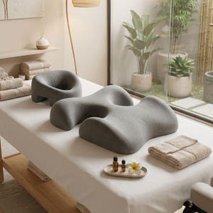 Peut inclure: Une scène de spa avec des oreillers de massage gris de différentes formes sur une table blanche. Une serviette beige pliée, des flacons d'huiles essentielles et une fleur sont sur un petit plateau. Des serviettes roulées et des plantes sont en arrière-plan.