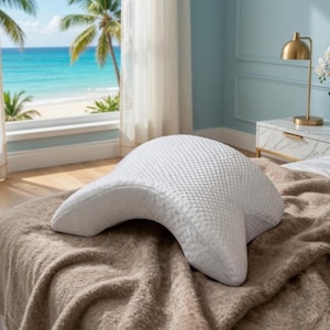 Puede incluir: Una almohada blanca contorneada con una superficie texturizada descansa sobre una cama cubierta con una manta marrón. La almohada tiene una forma única diseñada para brindar soporte. El fondo muestra una ventana con vista a la playa.