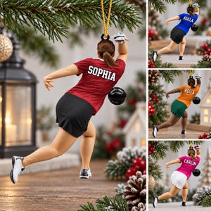 May include: A bowling-themed Christmas ornament featuring a figure in a red shirt with the name SOPHIA in white text, a black skirt, and white sneakers, holding a black bowling ball. The figure is suspended by a gold cord for hanging.