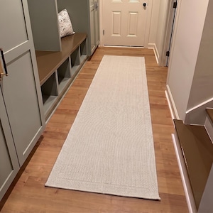 May include: A long, rectangular runner rug featuring a light gray, textured weave pattern with a subtle border detail. The rug is positioned lengthwise on a wooden floor, extending from the foreground toward a closed white door in the background.