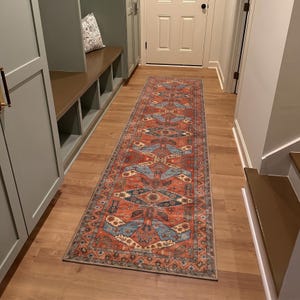 May include: A long, rectangular rug with an intricate pattern of blue, beige, and rust-coloured geometric shapes. The rug is placed on a wooden floor in a hallway, adding a touch of warmth and design to the space.