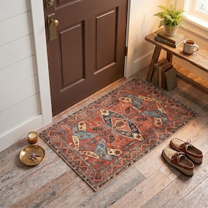 May include: A rectangular area rug featuring a complex pattern in shades of rust orange, navy blue, and cream. The rug is positioned on a light-colored wooden floor in front of a dark brown door, near a small wooden bench, a candle, and shoes.