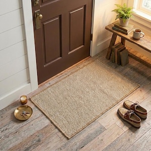 Puede incluir: Un felpudo rectangular tejido con un patrón de espiga en tonos beige y tostado. La alfombra está colocada sobre un suelo de madera clara frente a una puerta marrón oscuro, junto a un par de zapatos de cuero marrón y una bandeja dorada.