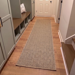 May include: A long, rectangular runner rug featuring a brown and tan herringbone pattern. The rug is positioned lengthwise on a light-coloured wooden floor in a hallway, extending from the foreground towards a white door in the background.