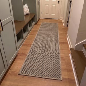 May include: A long, rectangular runner rug featuring a repeating herringbone pattern in shades of gray and white. The rug is positioned lengthwise on a light-toned wooden floor, extending from the foreground toward a closed white door in the distance.
