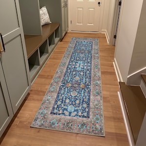 May include: A rectangular, patterned rug in a hallway. The rug features a dark blue central field with floral and geometric designs in shades of teal, pink, and beige. The border is a mix of teal, pink, and brown. The rug is placed on a wooden floor.