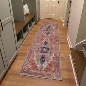 May include: A long rectangular runner rug featuring a faded terracotta red base with intricate patterns in navy blue, cream, and light blue. The design includes central diamond-shaped medallions and a decorative border, laid flat on a wooden floor.