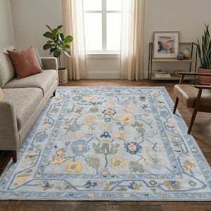 Op de afbeelding: Een rechthoekig vloerkleed met een lichtblauwe achtergrond en een herhalend bloemen- en rankenpatroon in tinten geel, groen, oranje en donkerblauw, omlijst door een decoratieve rand met bijpassende bloemmotieven en geometrische lijnen.