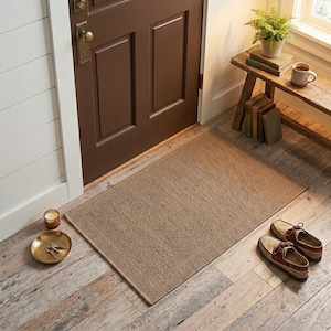 May include: A rectangular tan woven doormat with a textured surface and a border, positioned on a wooden floor in front of a dark brown door. The mat features a consistent weave pattern and sits flat, providing a neutral base for the entryway.