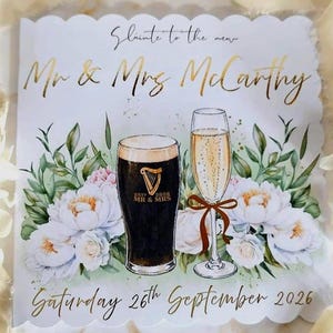 May include: A square wedding invitation card with scalloped edges featuring a dark stout beer and a champagne flute. Text reads Slainte to the new Mr & Mrs McCarthy and Saturday 26th September 2026. The design includes white floral accents.