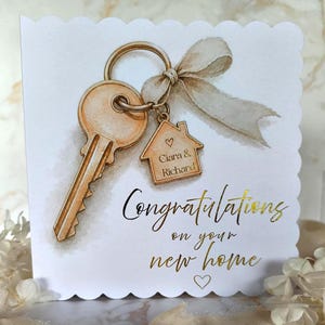 May include: A white greeting card with scalloped edges featuring a watercolour illustration of a gold key on a ring with a beige ribbon and a house-shaped charm engraved with the names Ciara & Richard. Gold foil text reads Congratulations on your new home.