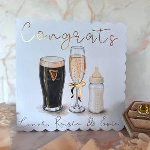 May include: A white scalloped greeting card featuring the gold foil text Congrats and the names Conor, Roisin & Evie. The illustration shows a pint of dark stout, a champagne flute with a gold ribbon, and a baby bottle filled with milk.