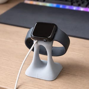 May include: A gray Apple Watch rests on a light gray charging stand. The watch has a black screen and a dark gray band. A white charging cable is plugged into the watch and the stand. The stand is on a wooden desk.