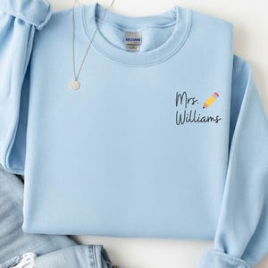 May include: A pale blue sweatshirt featuring the text "Mrs. Williams" and a pencil illustration. The sweatshirt is displayed with light wash denim jeans and a silver necklace. The sleeves are cuffed. The overall aesthetic is casual.