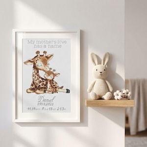 May include: A framed cross-stitch print featuring a mother giraffe and calf with the text My mother's love has a name, Daniel, 07.08.2025, 11:54pm, 7lbs 13oz, and 21.3in. Beside the frame sits a beige crocheted rabbit and small cotton stems.