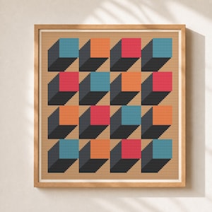 May include: A framed cross-stitch embroidery featuring a repeating geometric pattern of isometric cubes in shades of blue, orange, and red, set against a tan background. The design consists of sixteen cubes arranged in a four-by-four grid.