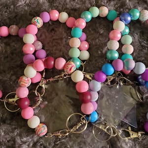May include: A collection of beaded wrist keychains in various colors, including pink, purple, teal, and white. Each keychain features a mix of solid and patterned beads, with gold-colored hardware and a clear plastic tag holder.