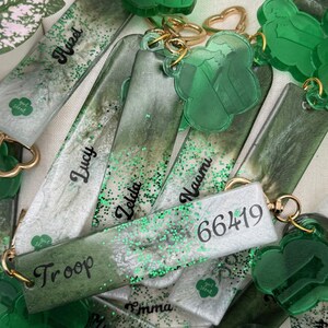 May include: A collection of green and white keychains with gold-colored hardware. Some keychains are rectangular with names like "Lucy" and "Zelda" in black script. Others are clover-shaped. The keychains have green glitter accents and the text "Girl Scout".