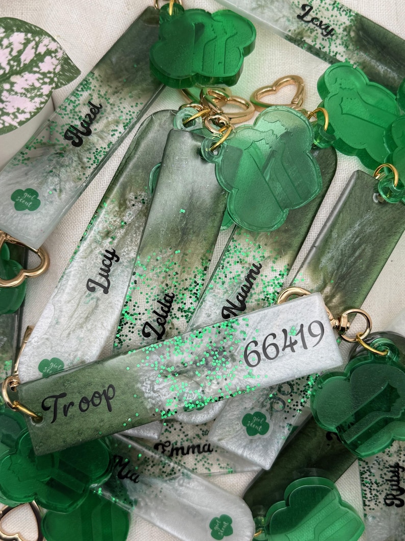 May include: A collection of green and white keychains with gold-colored hardware. Some keychains are rectangular with names like "Lucy" and "Zelda" in black script. Others are clover-shaped. The keychains have green glitter accents and the text "Girl Scout".