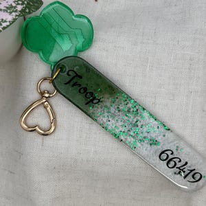 May include: A green and white keychain with the word "Troop" and the number "66419" printed on it. The keychain has a gold heart-shaped clasp and a green clover-shaped charm. The keychain is decorated with green glitter.