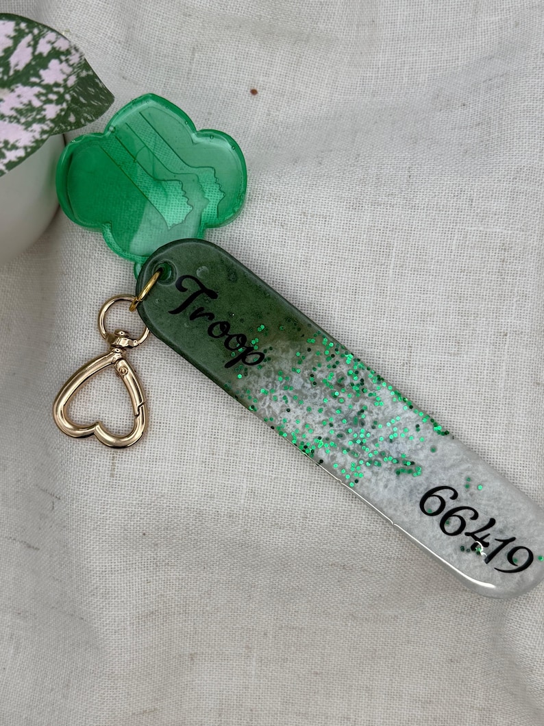 May include: A green and white keychain with the word "Troop" and the number "66419" printed on it. The keychain has a gold heart-shaped clasp and a green clover-shaped charm. The keychain is decorated with green glitter.