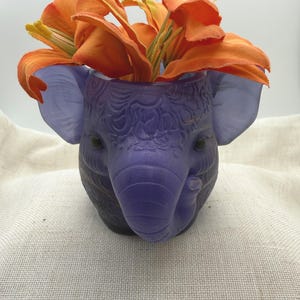 Può includere: Un vaso o contenitore traslucido di colore viola a forma di elefante, caratterizzato da intricati motivi in rilievo sulla superficie. L'elefante presenta grandi orecchie e una proboscide curva. Fiori di giglio artificiali arancioni sono disposti all'interno.