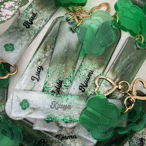 May include: A collection of personalized keychains and bookmarks. The keychains feature a green clover design and gold-colored hardware. The bookmarks are rectangular with glitter and names like "Hazel" and "Lucy" in black script. The color palette includes shades of green and white.