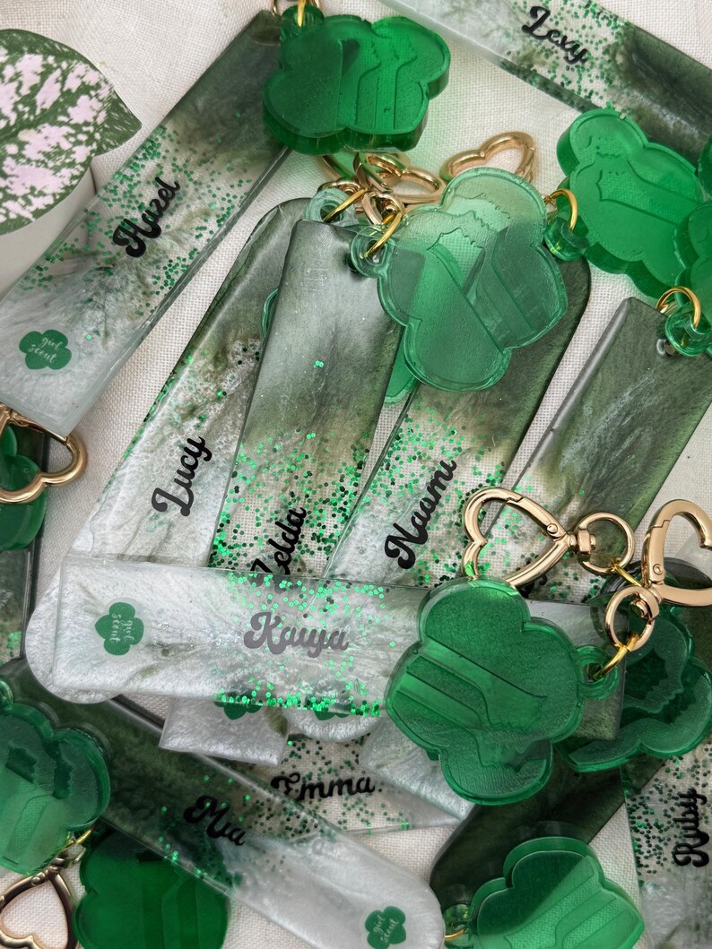 May include: A collection of personalized keychains and bookmarks. The keychains feature a green clover design and gold-colored hardware. The bookmarks are rectangular with glitter and names like "Hazel" and "Lucy" in black script. The color palette includes shades of green and white.