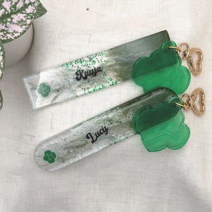 May include: Two rectangular keychains with green cloud charms. One keychain has the name "Kaiya" in cursive, the other has "Lucy". The keychains have a green and white glitter design with a green clover logo.