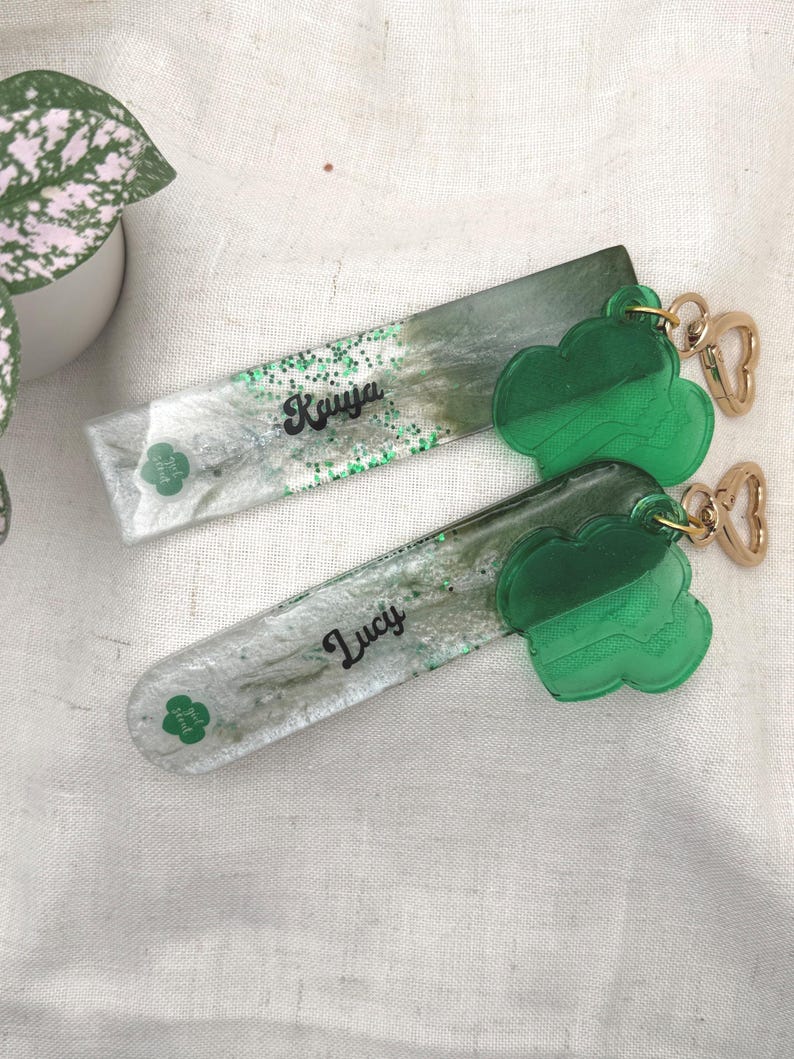 May include: Two rectangular keychains with green cloud charms. One keychain has the name "Kaiya" in cursive, the other has "Lucy". The keychains have a green and white glitter design with a green clover logo.