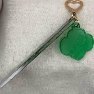 May include: A green Girl Scout cookie charm with a gold heart-shaped clasp and a clear, glitter-filled stick. The charm features a silhouette of a girl's face. The keychain is on a white fabric background.