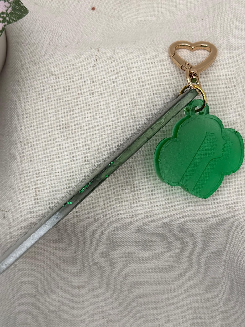 May include: A green Girl Scout cookie charm with a gold heart-shaped clasp and a clear, glitter-filled stick. The charm features a silhouette of a girl's face. The keychain is on a white fabric background.