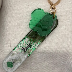 May include: A green and silver keychain with a heart-shaped clasp. The keychain features a green clover-shaped charm and a rectangular base with the name "Lexi" and the Girl Scout logo. The base is filled with glitter.