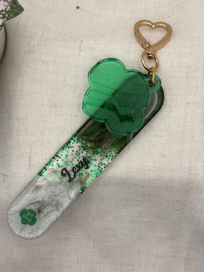 May include: A green and silver keychain with a heart-shaped clasp. The keychain features a green clover-shaped charm and a rectangular base with the name "Lexi" and the Girl Scout logo. The base is filled with glitter.
