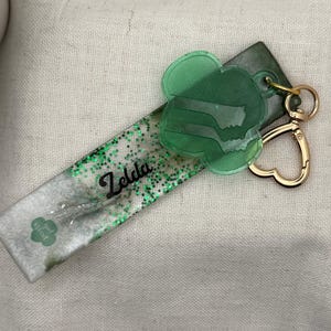 May include: A handmade keychain featuring a rectangular resin piece with green glitter and the name "Zelda" in black script. A green clover-shaped charm and a gold heart-shaped clasp complete the design. The keychain also has a small clover logo.