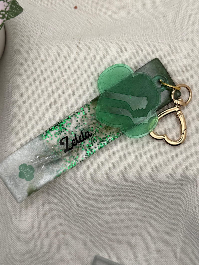 May include: A handmade keychain featuring a rectangular resin piece with green glitter and the name "Zelda" in black script. A green clover-shaped charm and a gold heart-shaped clasp complete the design. The keychain also has a small clover logo.