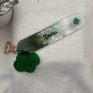May include: A personalized keychain with the name "Emma" in black script. The rectangular charm has a green and white design with green glitter and a green clover. A green clover charm and gold-colored clasp complete the accessory.