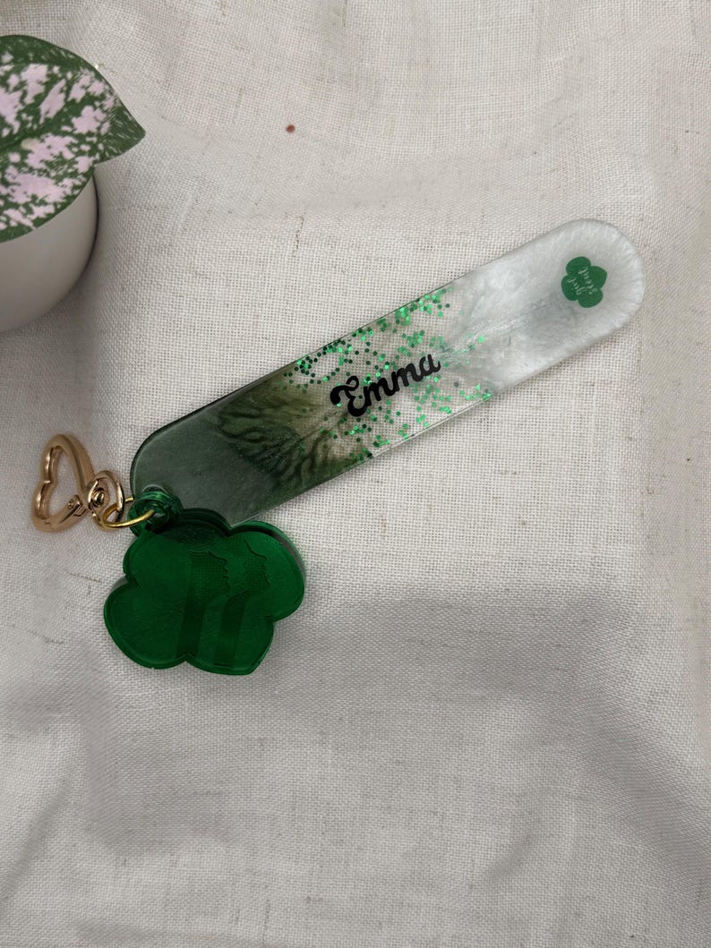 May include: A personalized keychain with the name "Emma" in black script. The rectangular charm has a green and white design with green glitter and a green clover. A green clover charm and gold-colored clasp complete the accessory.