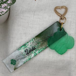 May include: A handmade keychain featuring a rectangular resin base with green glitter and the name "Zelda" in black script. A green clover-shaped charm and a gold heart-shaped clasp complete the design. The words "girl scout" are also visible.