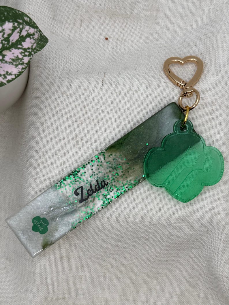 May include: A handmade keychain featuring a rectangular resin base with green glitter and the name "Zelda" in black script. A green clover-shaped charm and a gold heart-shaped clasp complete the design. The words "girl scout" are also visible.