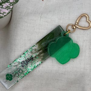 May include: A green and clear resin keychain with a gold-colored heart-shaped clasp. The keychain has a green clover-shaped charm and a rectangular section with the name "Abby" and the words "girl scout" in green glitter.