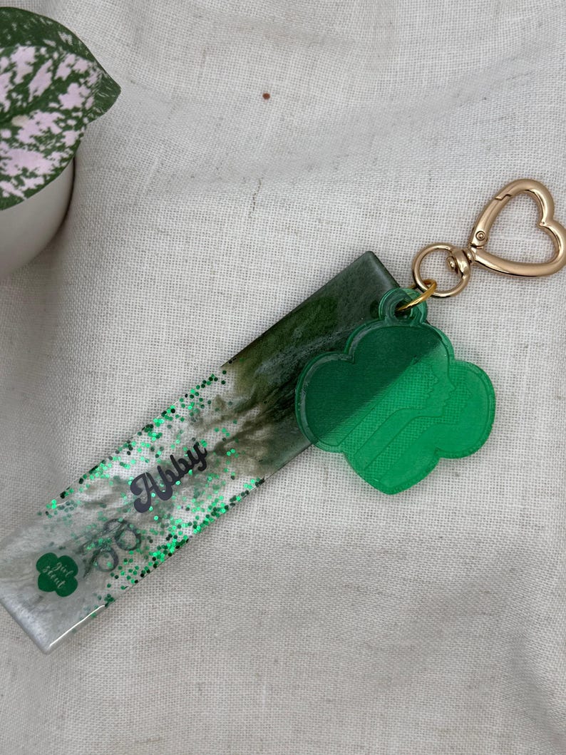 May include: A green and clear resin keychain with a gold-colored heart-shaped clasp. The keychain has a green clover-shaped charm and a rectangular section with the name "Abby" and the words "girl scout" in green glitter.