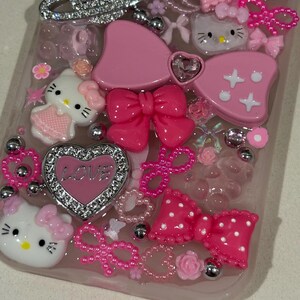 K&ouml;nnte beinhalten: Eine pinkfarbene Handyh&uuml;lle, verziert mit verschiedenen Verzierungen. Die H&uuml;lle zeigt Hello Kitty-Figuren, Schleifen, Herzen mit dem Wort "LOVE" und silberne Akzente. Das Design umfasst eine Mischung aus Texturen und Rosat&ouml;nen.