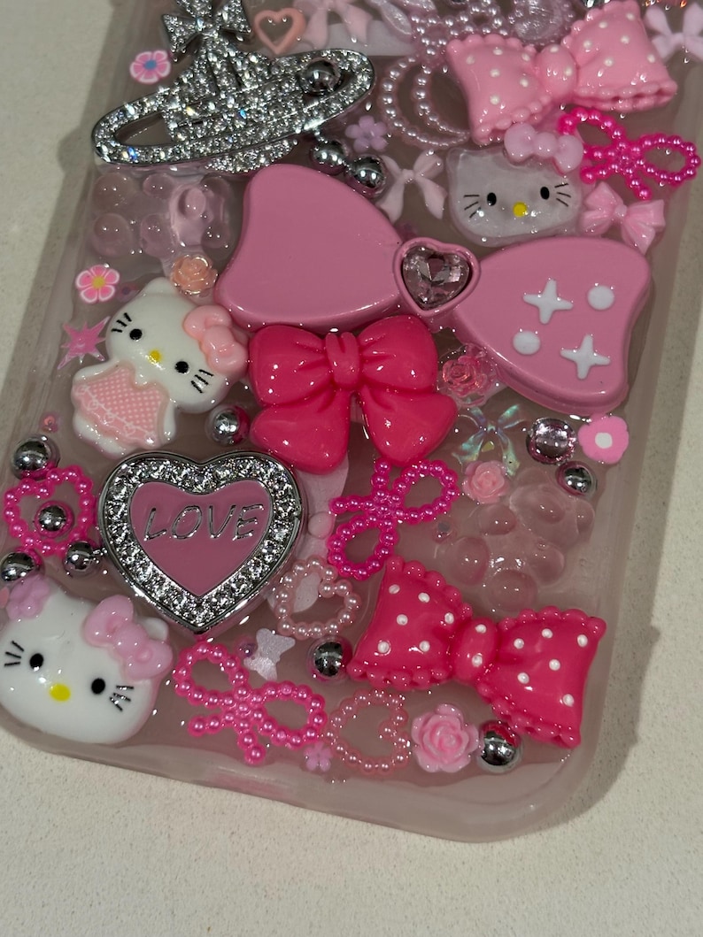 K&ouml;nnte beinhalten: Eine pinkfarbene Handyh&uuml;lle, verziert mit verschiedenen Verzierungen. Die H&uuml;lle zeigt Hello Kitty-Figuren, Schleifen, Herzen mit dem Wort "LOVE" und silberne Akzente. Das Design umfasst eine Mischung aus Texturen und Rosat&ouml;nen.