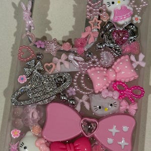 K&ouml;nnte beinhalten: Eine transparente Handyh&uuml;lle, verziert mit einer Vielzahl von rosa Verzierungen. Enth&auml;lt Hello Kitty-Figuren, Schleifen, Herzen, Sterne und ein glitzerndes silbernes Emblem. Die H&uuml;lle ist mit dekorativen Elementen in verschiedenen Texturen und Gr&ouml;&szlig;en verziert.